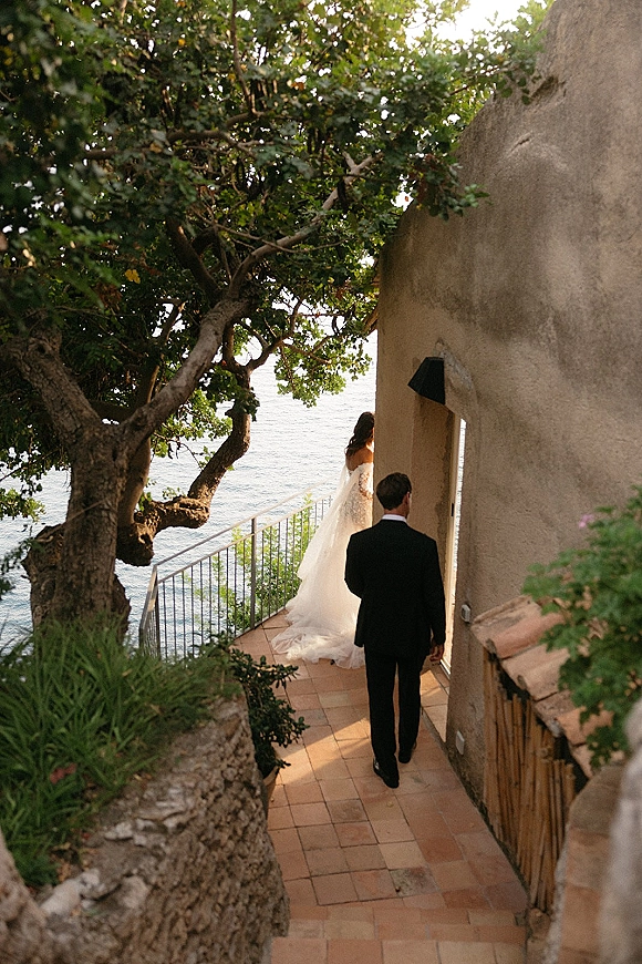 Wedding first look as bride in a veil and long train walks toward groom in a black tuxedo on a seaside terrace walkway with ocean view