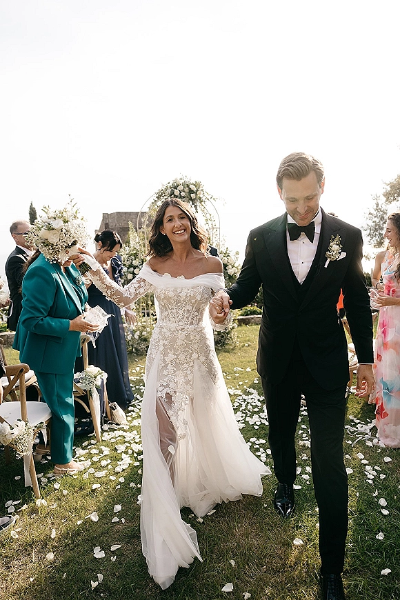 Wedding recessional as bride and groom walk the aisle holding hands, bouquet in hand, past rose petals under a bright lawn sky