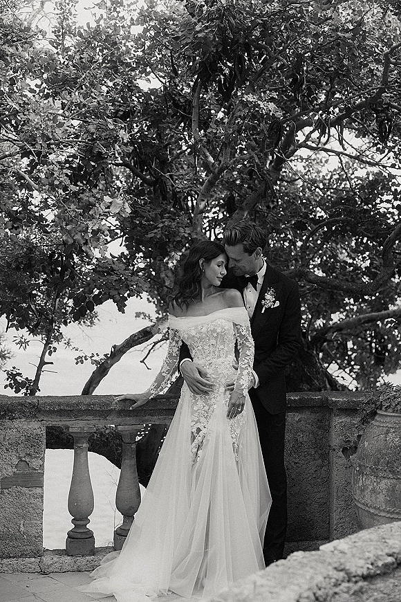 Couple portrait in a black and white wedding portrait style, bride in off-shoulder lace gown embraced by groom in tux by stone balustrade with trees behind