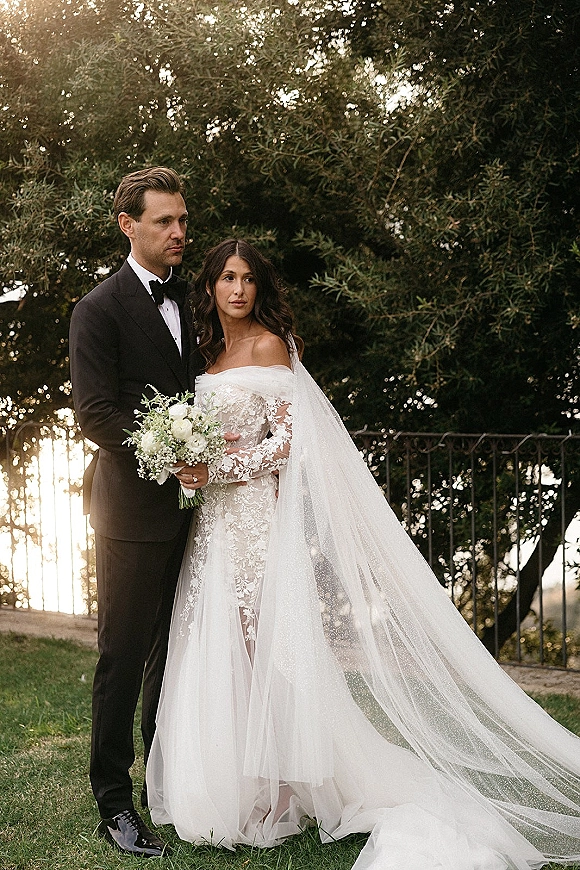 Couple portrait of bride in lace gown and long veil with groom in black tuxedo, holding a white-and-green bouquet at sunset lawn by iron fence