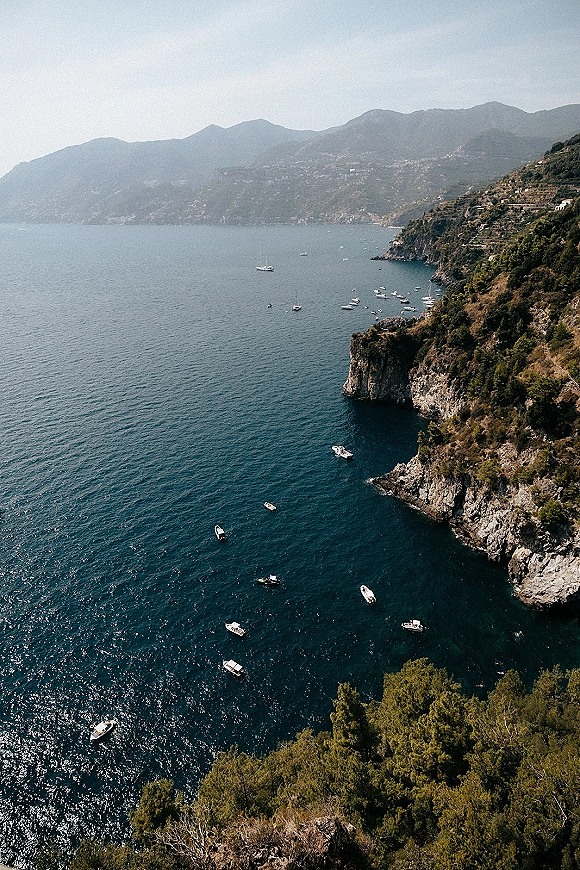 Coastal landscape with boats offshore, featuring rocky cliffs and turquoise ocean water beneath hills, mountains, and clear sky