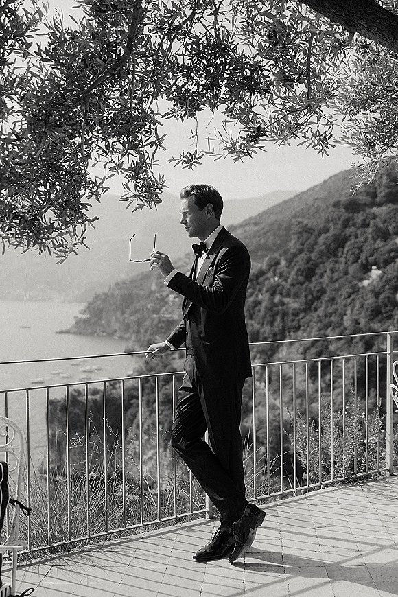 Groom portrait in a tuxedo, wearing sunglasses and leaning on a terrace railing with coastline, boats, and sea in the distance