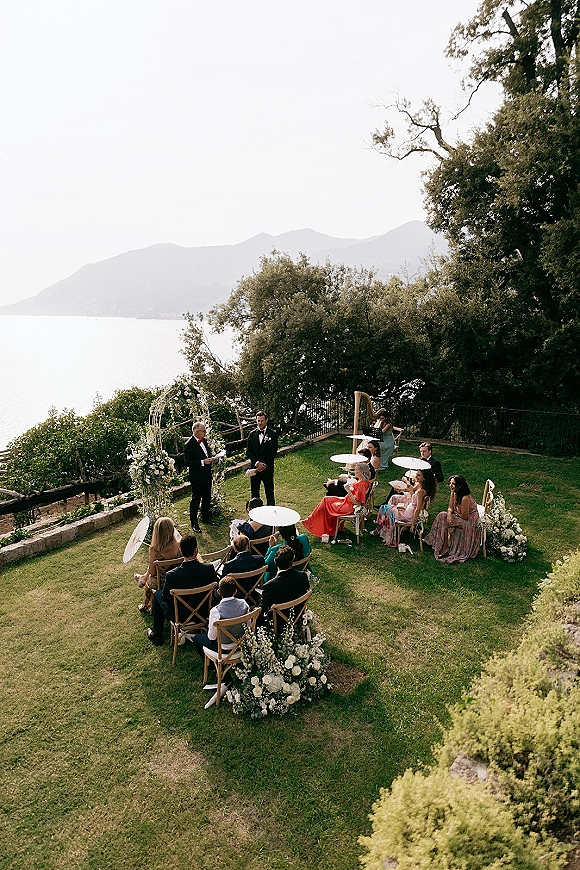 Outdoor wedding ceremony on a lawn with guests under white parasols facing a floral arch, harp and mics, with lake and mountains beyond