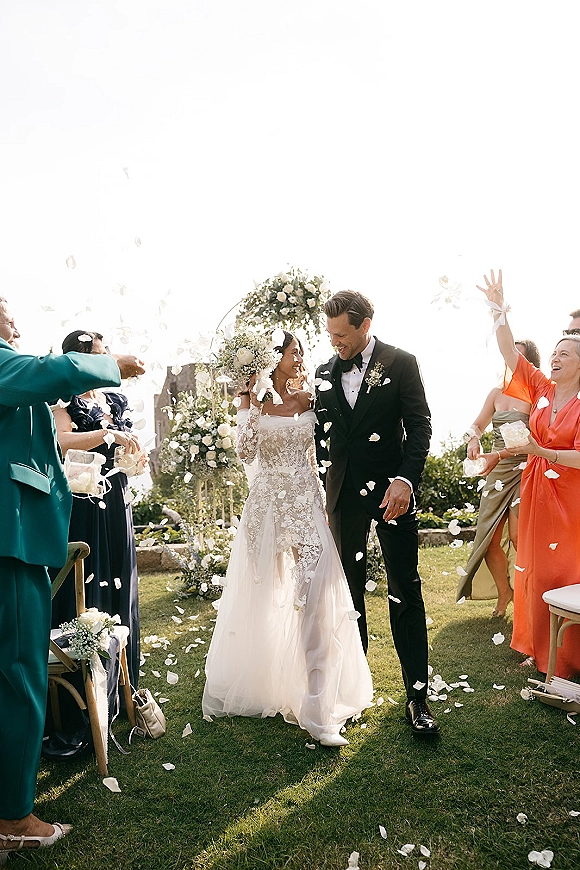 Wedding recessional as bride and groom walk down the aisle while guests toss flower petals on a sunlit garden lawn under a floral arch