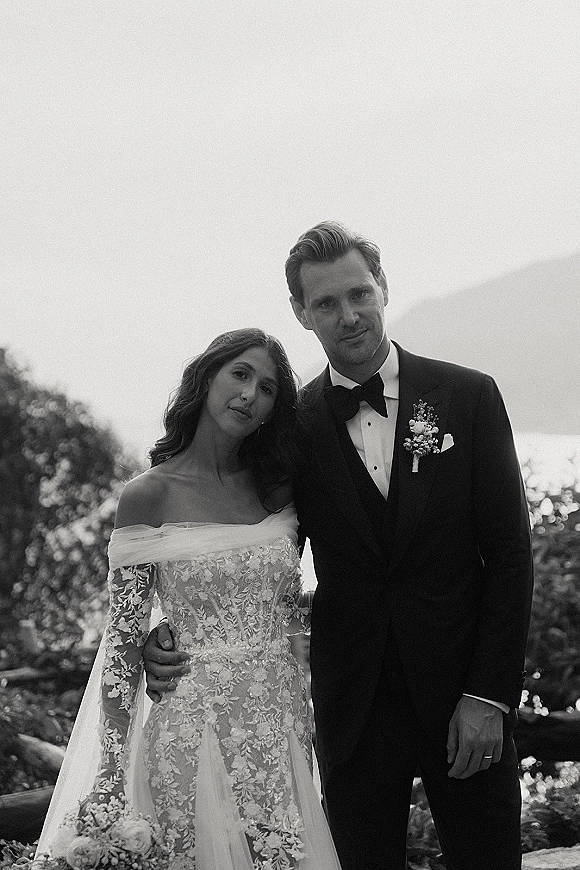 Couple portrait in a black and white wedding portrait style, groom with arm around bride in lace long sleeves by a garden fence
