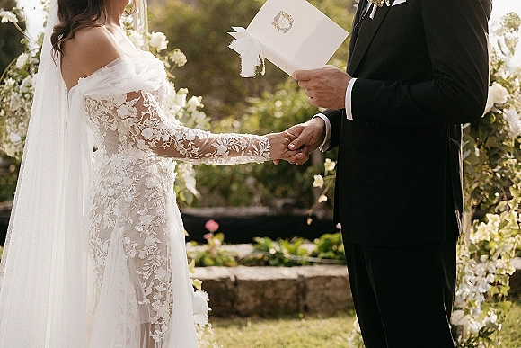 Wedding vows as bride and groom hold hands while he reads from a ribbon vow book at a sunlit garden ceremony on stone steps
