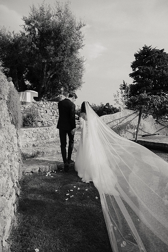 Couple portrait of bride and groom from behind holding hands on stone steps, her long veil trailing over rose petals in a garden setting