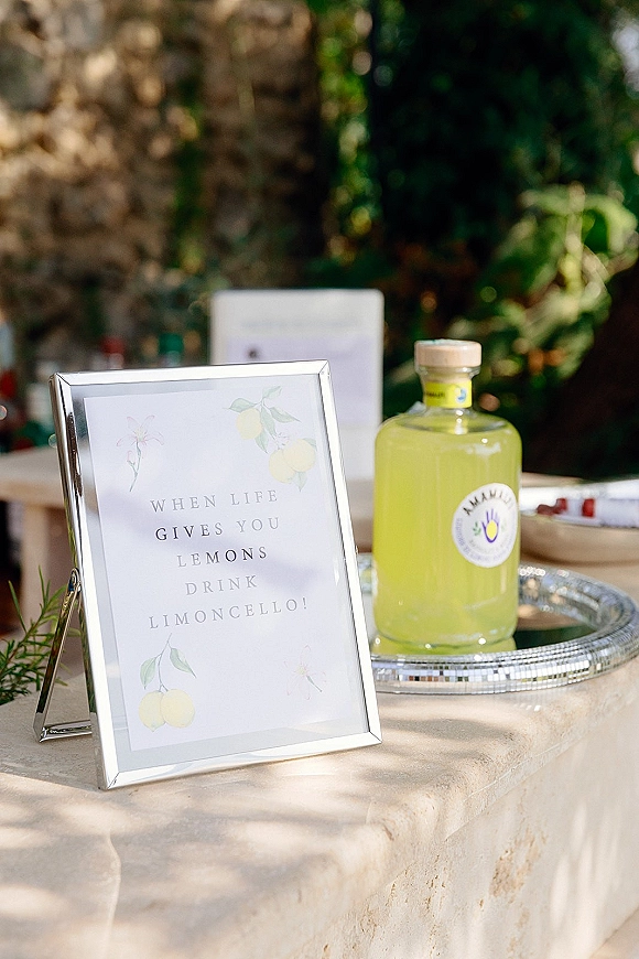 Wedding bar signage in a silver frame, signature drink sign beside a limoncello bottle on a tray at an outdoor garden table by a stone wall