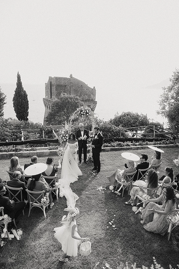 Wedding ceremony with bride in veil walking the petal-strewn aisle toward a floral arch on a seaside lawn with stone tower and parasols