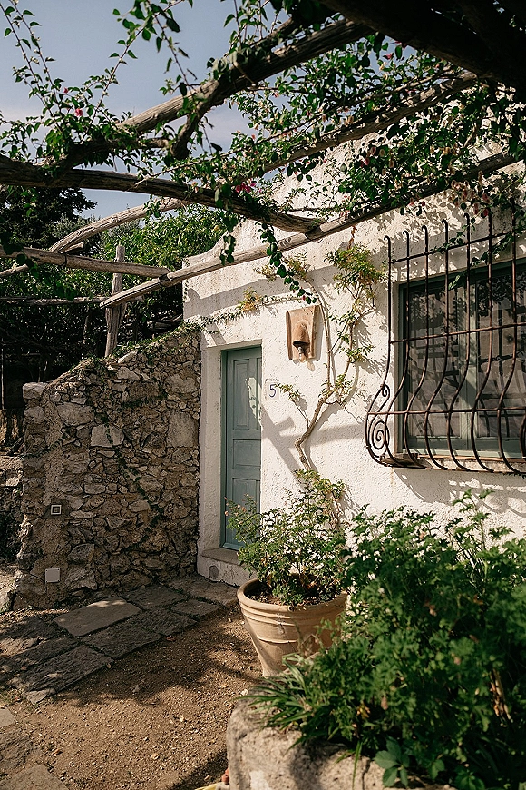 Outdoor venue exterior with a vine-covered pergola framing a painted door, beside a white stucco wall and stone path entry