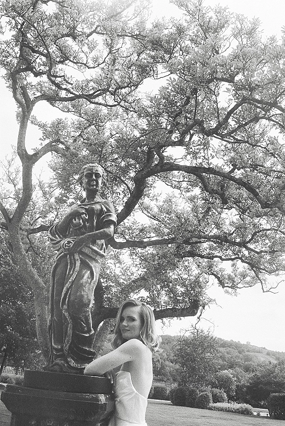 Bridal portrait of a bride by statue in a strapless wedding dress, leaning in side profile in a garden with trees and hills beyond