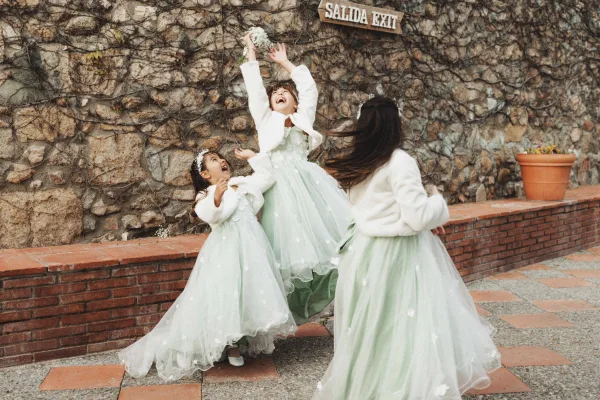 Flower girl moment as two girls in mint tulle dresses toss flower petals by a stone wall, laughing mid-step in winter jackets