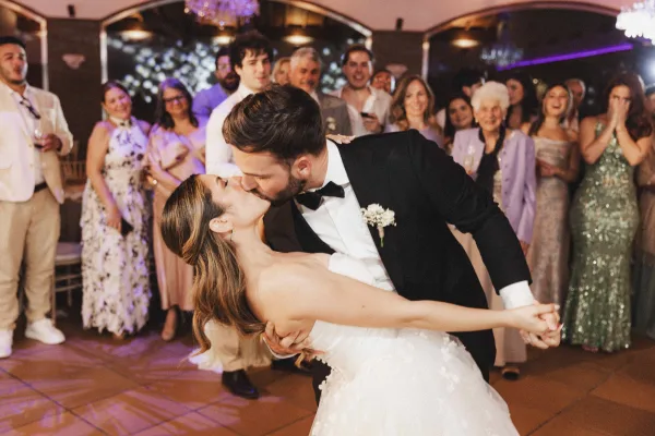 Wedding first dance dip as bride in strapless gown and groom in tuxedo share a kiss on dance floor with purple uplighting and guests watching
