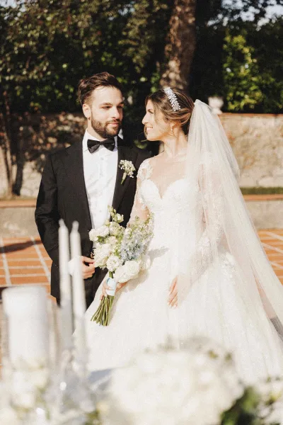 Couple portrait of bride in lace gown with veil and bouquet beside groom in black tuxedo on a terracotta garden terrace with candles