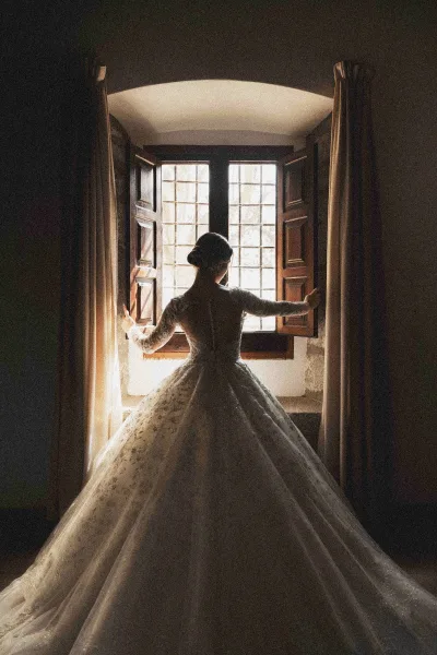 Bridal portrait of a bride at window, seen from behind holding curtains to show button-back lace long-sleeve gown in soft natural light