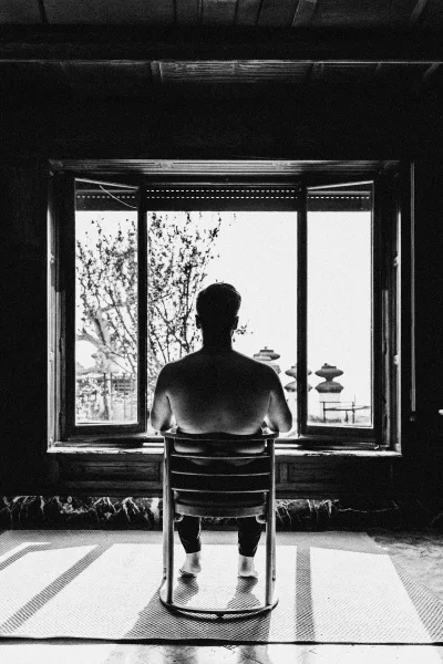 Groom getting ready in socks, sitting in a chair by a window, back view in soft indoor light with rooftops outside
