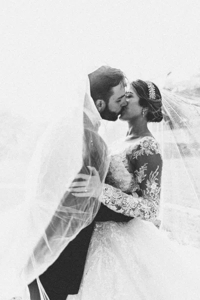 Wedding kiss portrait of bride and groom kissing under a windblown veil, lace long-sleeve gown and dark suit against a bright sky