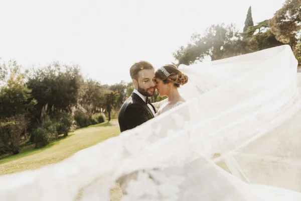 Couple portrait of bride and groom portrait in close embrace, bride veil blowing in sunlight on a garden lawn with trees