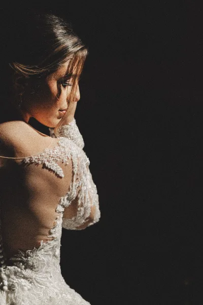 Bridal portrait of a bride in profile looking down, wearing an off-the-shoulder lace gown with choker necklace against a dark backdrop