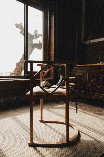Wedding shoes in window light on a chair with a ribbon accent, bouquet nearby in a vintage interior with wood paneling and rug