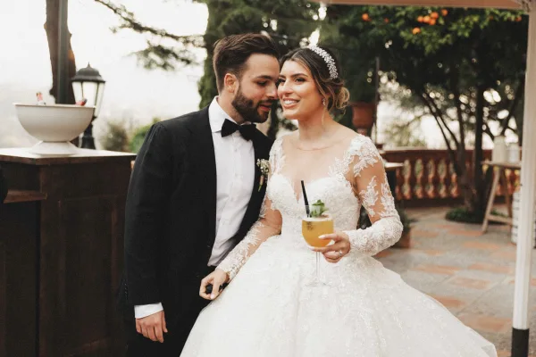 Couple portrait of bride and groom portrait on a dusk terrace, groom leaning in as bride holds a cocktail under string lights with veil