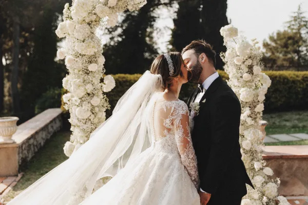 Wedding kiss portrait of bride and groom kiss under a white floral arch, her long veil and lace dress in a garden setting