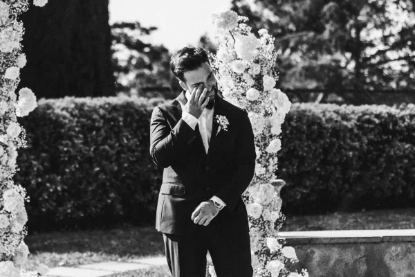 Groom portrait of a tuxedo groom wiping tears under a white floral arch, bow tie and boutonniere set against garden hedges and stone wall