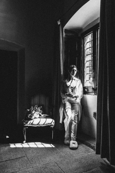 Bridal portrait in a lace robe by window light in a moody dark room, holding a champagne flute beside bouquet on bench