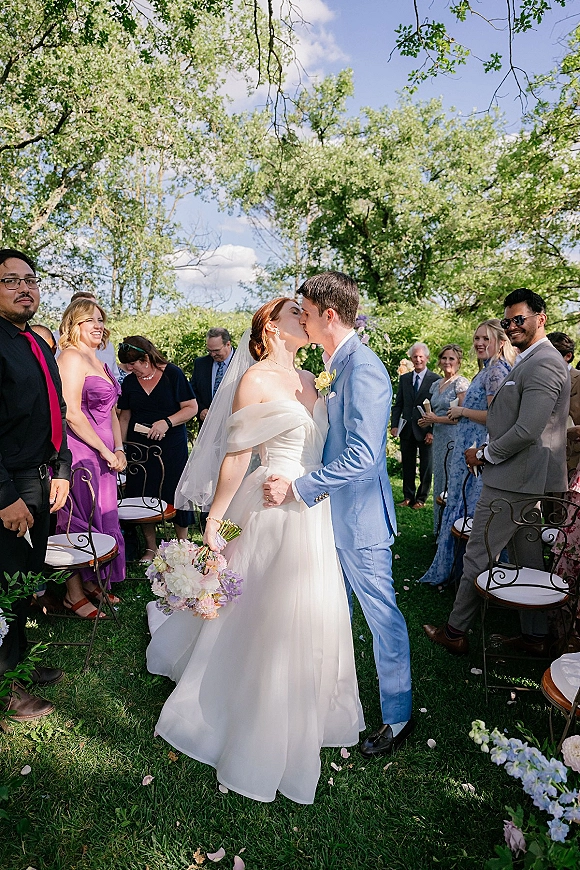 Wedding kiss as newlyweds share a ceremony kiss moment in the garden aisle, bride in veil with bouquet and groom in light blue suit, petals underfoot