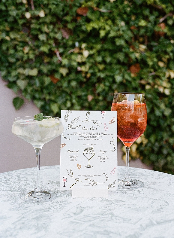 Wedding bar menu printed on a stand beside spritz cocktails with ice and citrus garnish on a table, set against a greenery wall patio backdrop
