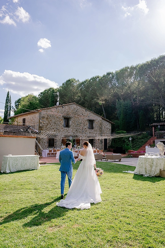 Couple portrait of bride and groom walking away holding hands, her long veil and bouquet trailing by a stone villa courtyard reception