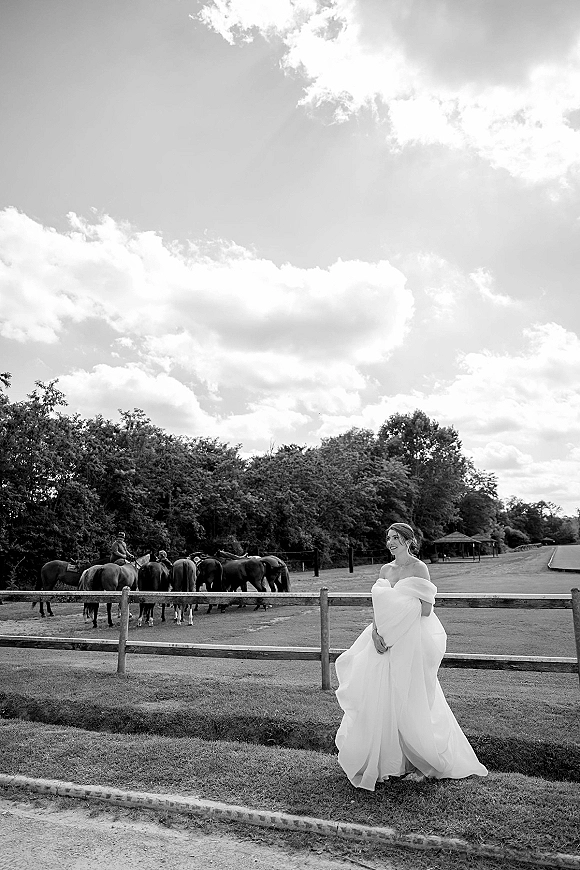 Bridal portrait of a bride in an off the shoulder wedding dress holding her skirt by a wood fence, with horses in a pasture under cloudy sky