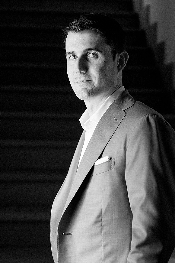 Groom portrait in a light suit jacket with dress shirt and pocket square, standing by an interior staircase in soft shadows