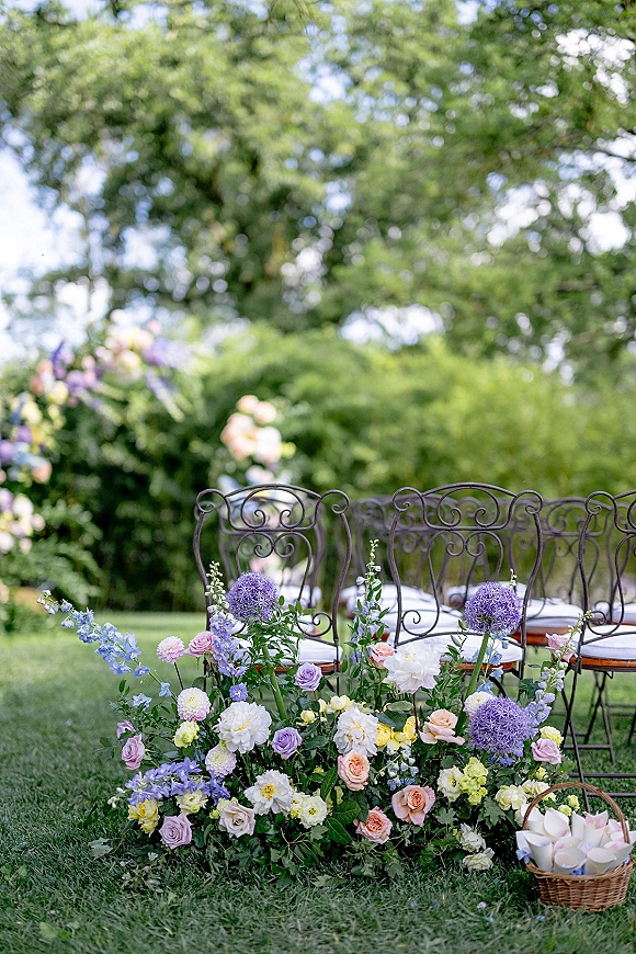 Ceremony aisle decor with outdoor ceremony flowers in pastel arrangements of purple allium, roses and delphinium beside garden chairs on lawn