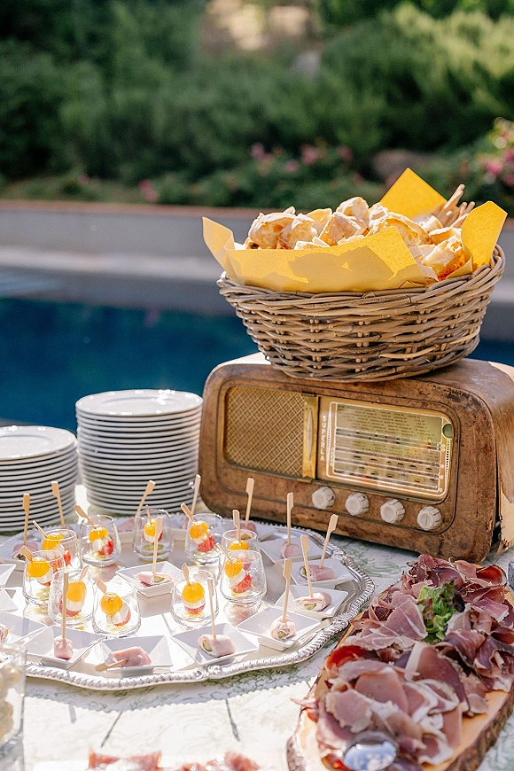 Wedding appetizer table with cocktail hour appetizers on silver trays, skewers, prosciutto and bread baskets beside a pool in garden greenery