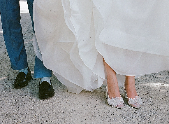 Wedding shoes with bridal heels close up, floral appliqué peeking under the dress hem beside groom black dress shoes on gravel outdoors