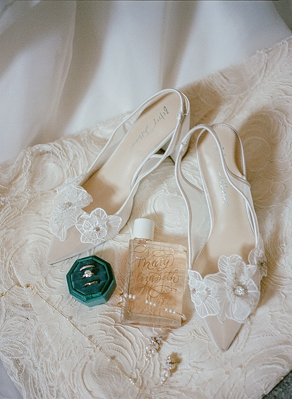 Bridal shoe flatlay of white bridal heels with lace appliques and pearl accents, paired with rings on lace wedding dress fabric