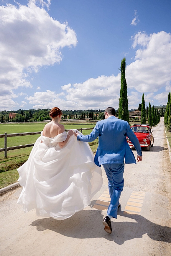 Couple portrait of bride and groom walking away on a country road, she lifts her gown train toward a red vintage car under open sky