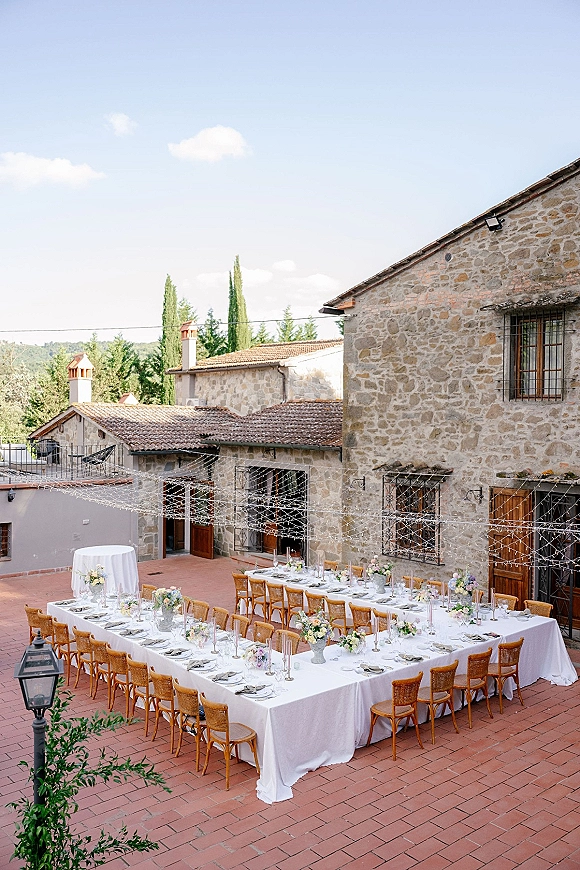 Outdoor reception tablescape in a courtyard wedding reception with long U-shaped banquet tables, pastel florals, taper candles, and overhead string lights