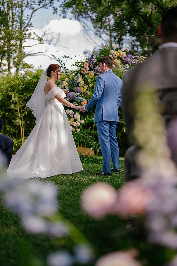 Wedding vows during an outdoor wedding ceremony as bride and groom hold hands beneath a pastel floral arch on a garden lawn with guests watching
