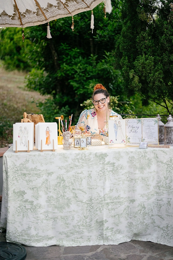 Wedding live painter table display with easels, illustration cards, and paintbrushes set on an outdoor patio amid garden greenery and trees