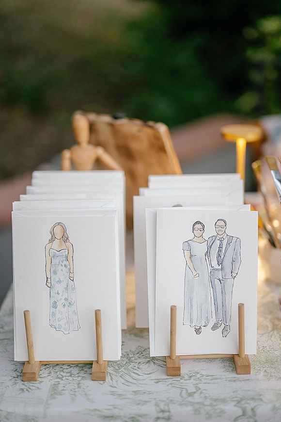 Escort cards arranged as a wedding escort cards display with illustrated portraits in wooden card stands on a tablecloth, greenery behind