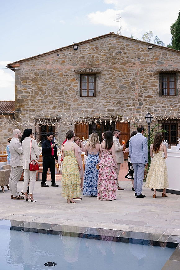 Wedding cocktail hour with guests mingling around an outdoor wedding bar, cocktails in hand, on a stone villa patio by the pool under string lights