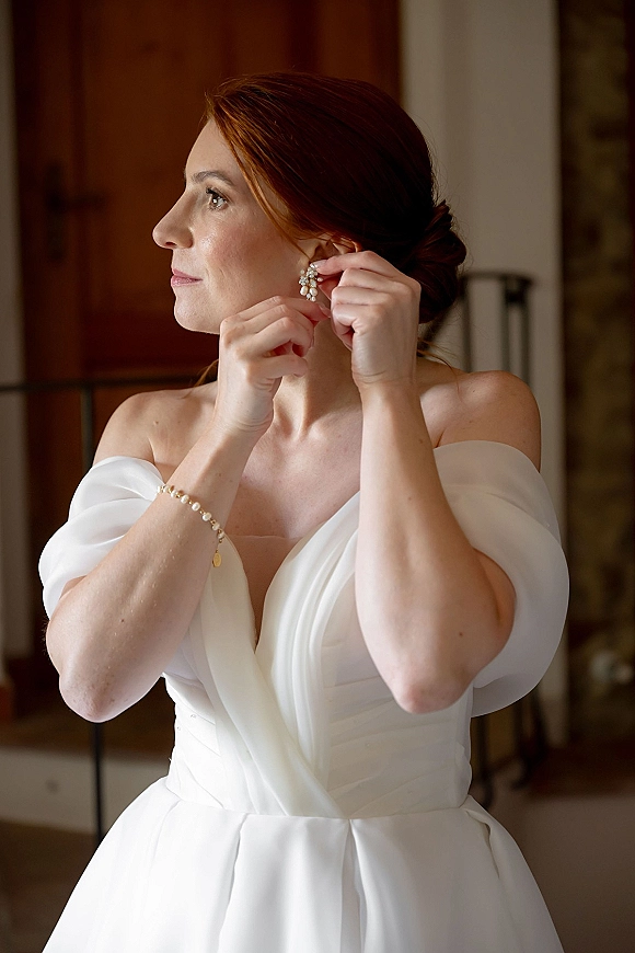 Bride getting ready, adjusting pearl drop wedding earrings in an indoor room, wearing an off the shoulder dress with hair in a bun updo.