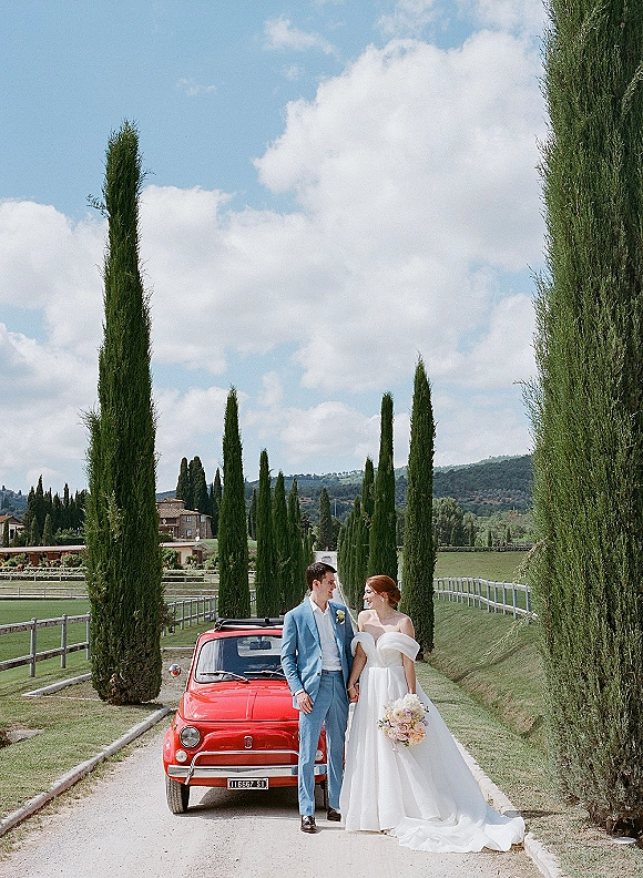 Couple portrait of bride and groom by car, leaning on a red vintage Fiat on a tree-lined country road with cypress trees under clouds