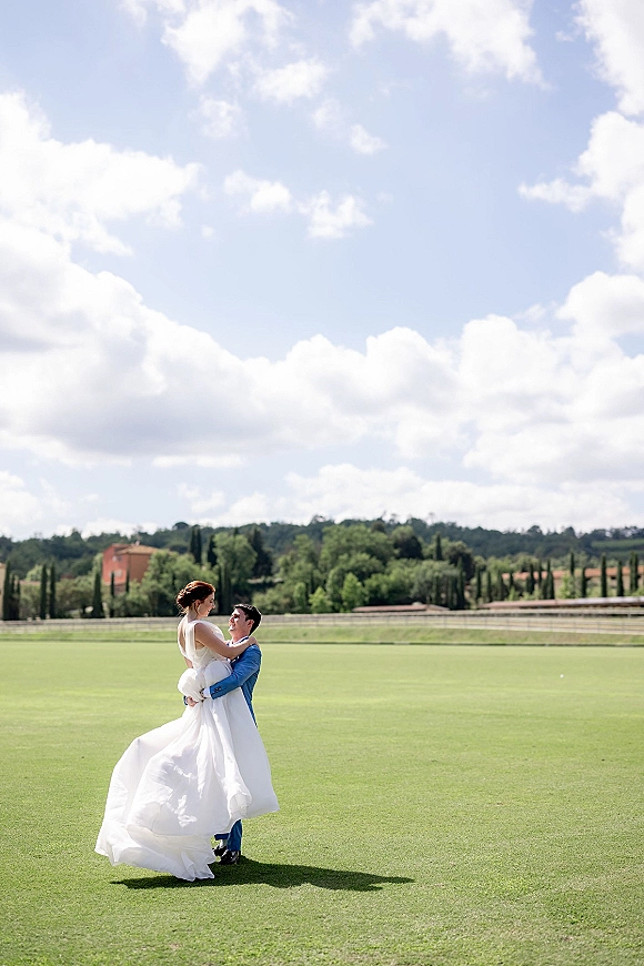 Couple portrait of groom in a blue suit lifting his bride, her veil and flowing wedding dress against a cloudy sky on a green lawn