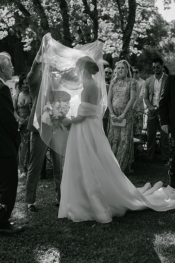 Wedding kiss portrait of bride and groom under a veil, holding a white bouquet as guests watch in a sunlit garden ceremony