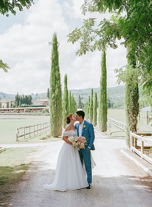 Wedding kiss portrait of bride and groom kissing, she holds a bouquet in an off-the-shoulder dress on a tree-lined driveway under clouds