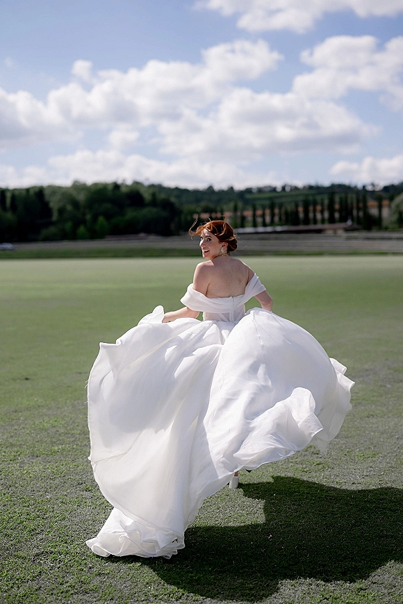 Bridal portrait of a bride running and looking back, her off-the-shoulder gown billowing on a grass field under blue sky