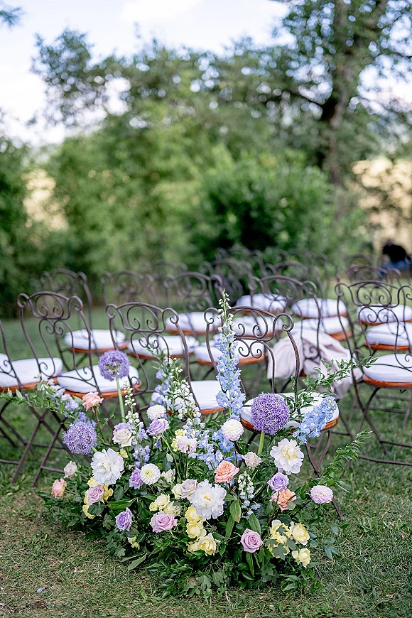 Ceremony aisle decor with wrought iron chairs and white cushions beside a low rose, delphinium and allium floral ground arrangement on a lawn
